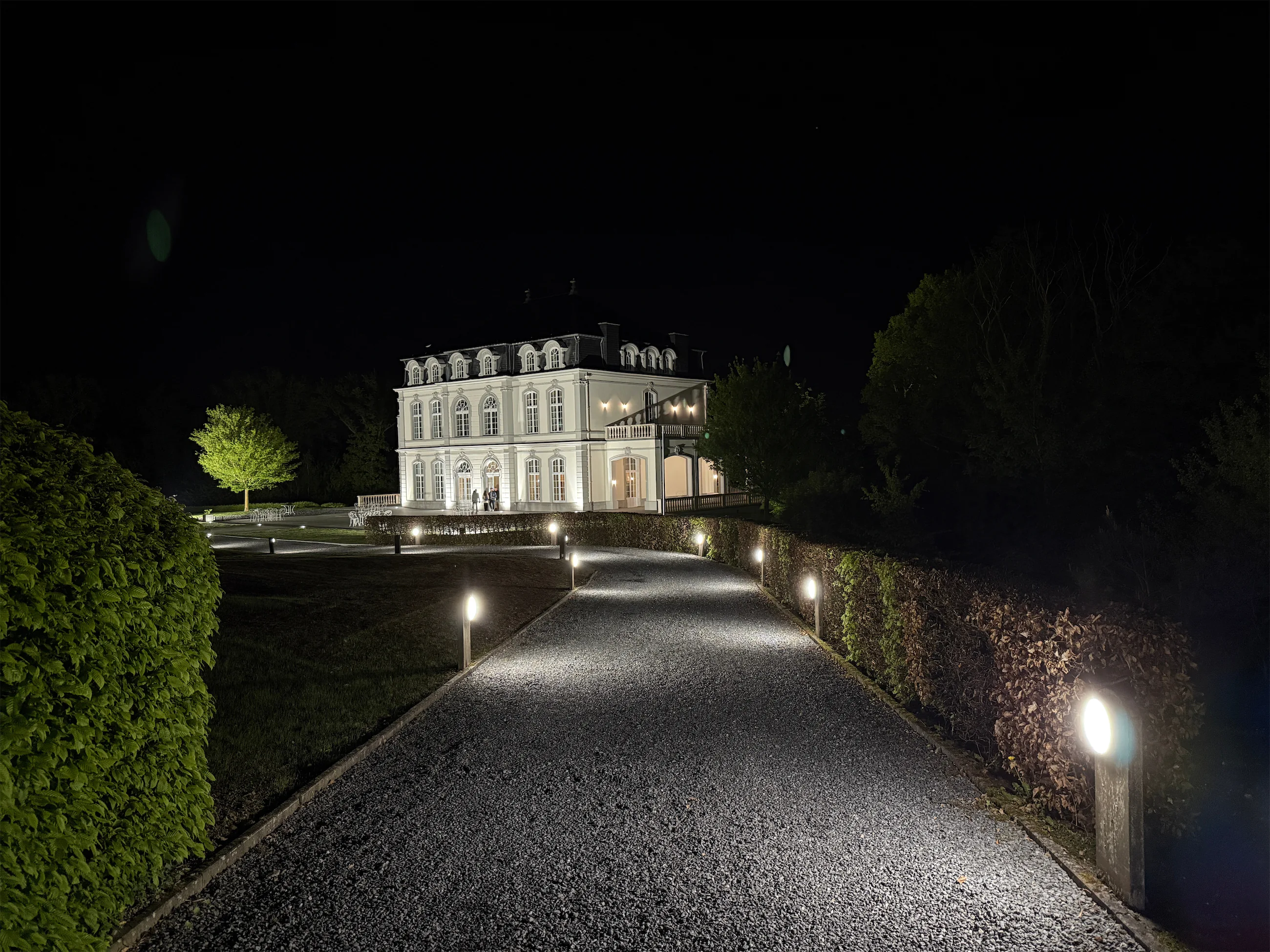 Château blanc illuminé la nuit, allée bordée de haies et lanternes
