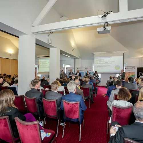 Salle de conférence avec audience assise sur chaises rouges face à un écran de projection