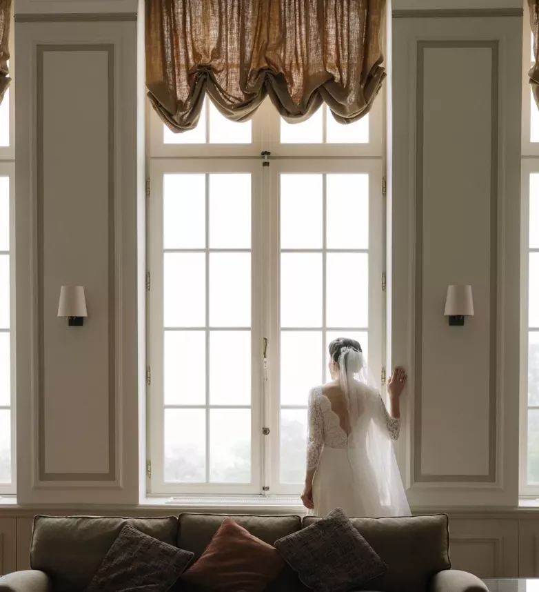 Mariée en robe blanche debout devant une grande fenêtre lumineuse dans une pièce élégante