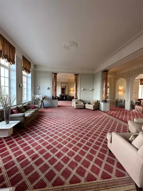 Grand salon avec moquette à motifs écossais rouge, canapés beiges et hautes fenêtres lumineuses