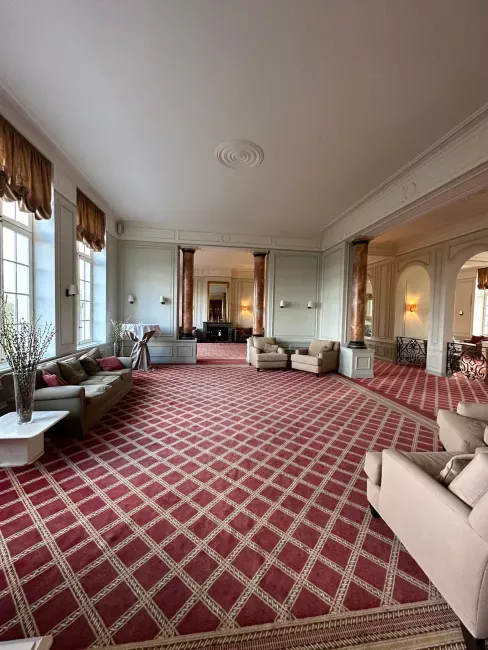 Grand salon avec moquette rouge à motifs, canapés beiges et grandes fenêtres lumineuses