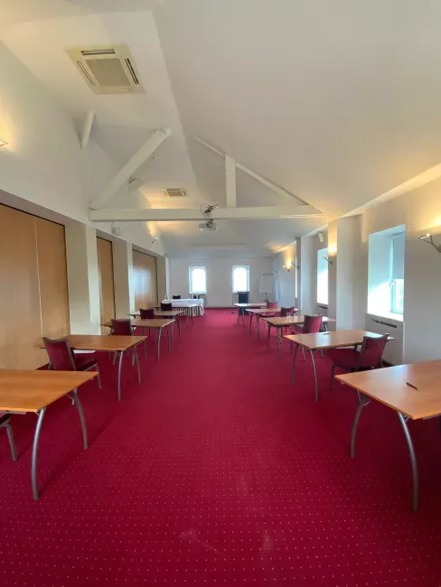 Salle de réunion avec moquette rouge, tables et chaises disposées en rangs, plafond blanc avec poutres